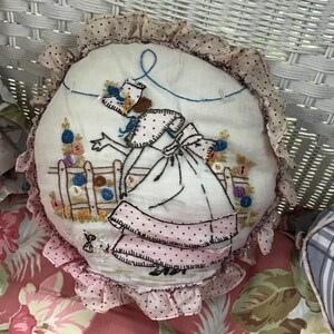 K&ouml;nnte beinhalten: Ein rundes, wei&szlig;es Kissen mit einem rosa-wei&szlig;en R&uuml;schenbesatz. Das Kissen ist mit einer Frau in einem rosa Kleid und einem blauen Hut bestickt, die in einem Garten mit einem Zaun und Blumen steht.