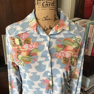 May include: A button-down shirt with a light blue background and a white pattern. The shirt features a colorful floral design with orange, pink, and green accents. The long-sleeved shirt is displayed on a mannequin.