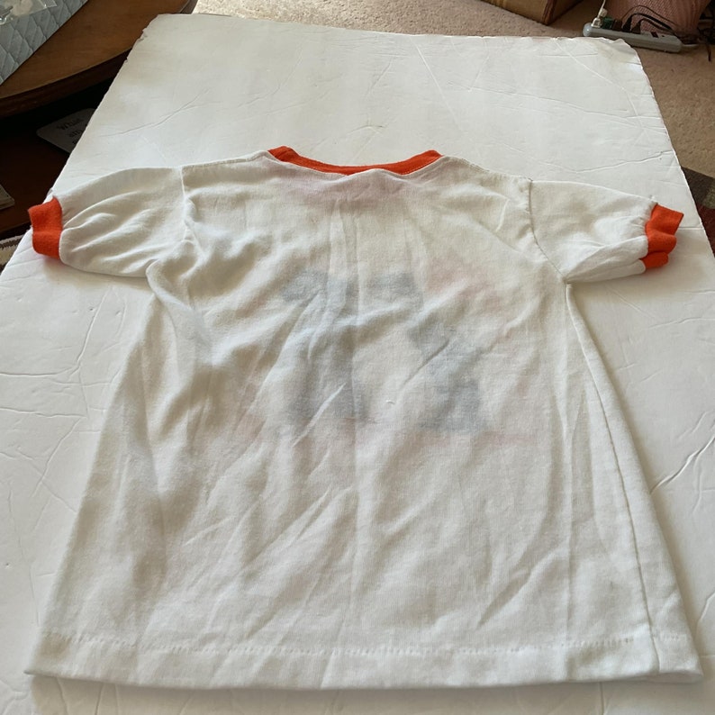May include: White t-shirt with orange trim around the neck and sleeves. The shirt has a faded graphic on the back.