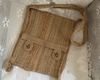 May include: A woven jute shoulder bag in a natural tan color. The bag features a flap closure with two decorative buttons and a long strap. The bag is rectangular with a textured surface and a simple, rustic design.