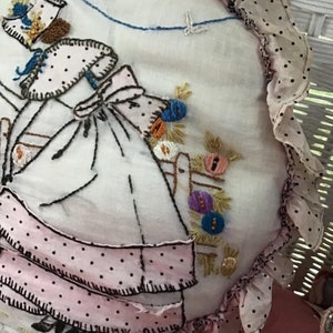 K&ouml;nnte beinhalten: Ein handgefertigtes besticktes Kissen mit einem rosa-wei&szlig;en Tupfenmuster. Das Kissen zeigt ein M&auml;dchen in einem rosa Kleid und einem blauen Band, das einen Blumenkorb h&auml;lt.