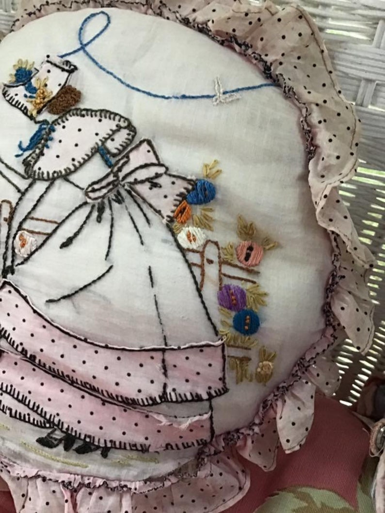 K&ouml;nnte beinhalten: Ein handgefertigtes besticktes Kissen mit einem rosa-wei&szlig;en Tupfenmuster. Das Kissen zeigt ein M&auml;dchen in einem rosa Kleid und einem blauen Band, das einen Blumenkorb h&auml;lt.