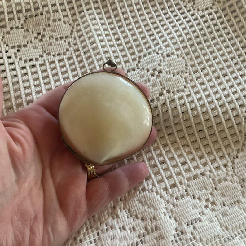 May include: A round, off-white compact with a gold-colored metal frame. The compact has a small loop at the top for hanging. The surface of the compact is smooth and appears to be made of a solid material.
