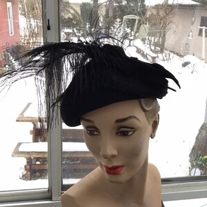 Puede incluir: Un gorro de fieltro negro con un largo adorno de plumas negras. El gorro est&aacute; en una cabeza de maniqu&iacute;.