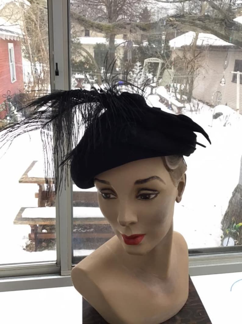 Puede incluir: Un gorro de fieltro negro con un largo adorno de plumas negras. El gorro est&aacute; en una cabeza de maniqu&iacute;.