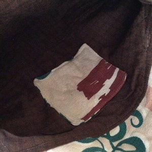 May include: A brown fabric tote bag with a small pocket on the inside. The pocket is made of a beige fabric with brown and white abstract designs. The bag has a green and white floral pattern on the outside.
