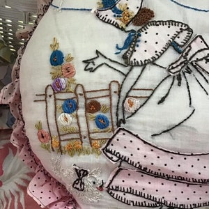 K&ouml;nnte beinhalten: Ein wei&szlig;es besticktes Kissen mit einem rosa und wei&szlig;en Volant mit wei&szlig;en Punkten. Die Stickerei zeigt eine Frau in einem rosa Kleid mit schwarzen Punkten, die einen blauen Vogel h&auml;lt. Die Frau steht neben einem Zaun mit einem Blumengarten.