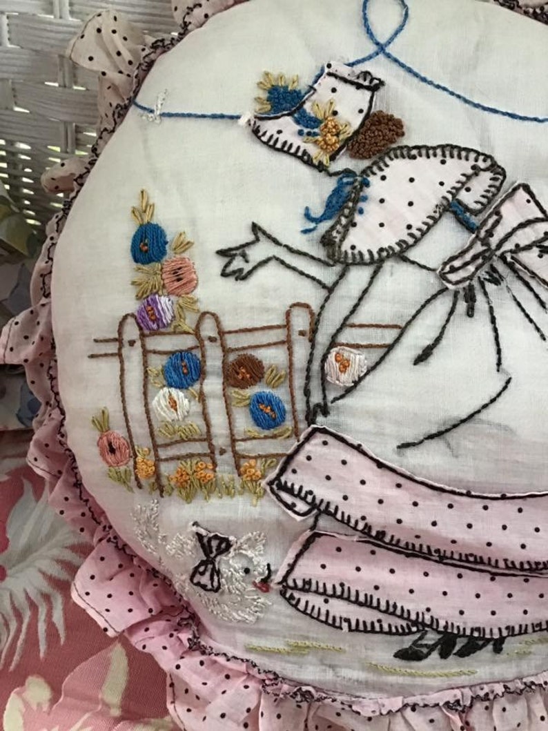 K&ouml;nnte beinhalten: Ein wei&szlig;es besticktes Kissen mit einem rosa und wei&szlig;en Volant mit wei&szlig;en Punkten. Die Stickerei zeigt eine Frau in einem rosa Kleid mit schwarzen Punkten, die einen blauen Vogel h&auml;lt. Die Frau steht neben einem Zaun mit einem Blumengarten.