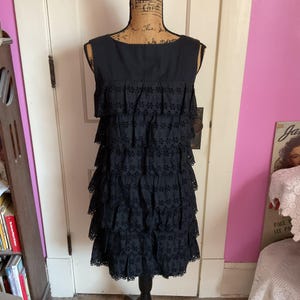 May include: A sleeveless, black dress with a tiered, eyelet skirt. The dress is displayed on a mannequin. The top of the dress is a solid black fabric, while the skirt features multiple layers of eyelet fabric with a scalloped hem.