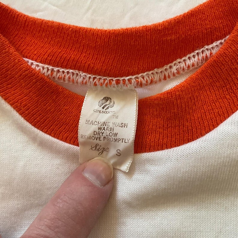 May include: White t-shirt with an orange trim around the neck. The tag says "Girl Scouts" and "Machine Wash Warm, Dry Low, Remove Promptly" and "Size S".