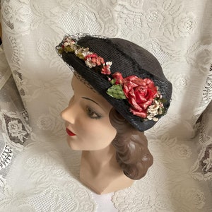 May include: A black straw hat with a wide brim and a floral embellishment. The flowers are red, pink, white, and green. The hat is trimmed with black netting.