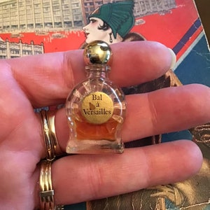 Puede incluir: Un frasco de perfume en miniatura de vidrio transparente con tap&oacute;n dorado y una etiqueta redonda con el texto Bal &agrave; Versailles. El frasco contiene un l&iacute;quido de color &aacute;mbar y tiene un cuerpo redondeado con base plana de dise&ntilde;o cl&aacute;sico.