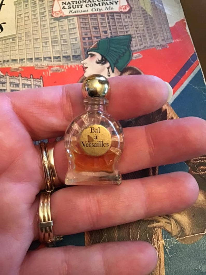 Puede incluir: Un frasco de perfume en miniatura de vidrio transparente con tap&oacute;n dorado y una etiqueta redonda con el texto Bal &agrave; Versailles. El frasco contiene un l&iacute;quido de color &aacute;mbar y tiene un cuerpo redondeado con base plana de dise&ntilde;o cl&aacute;sico.