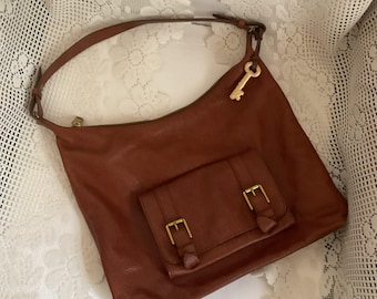 Puede incluir: Un bolso de hombro de cuero marrón con un bolsillo frontal a juego asegurado por dos hebillas doradas. El bolso tiene una correa larga y un dije dorado en forma de llave. El bolso está sobre un fondo de encaje blanco.