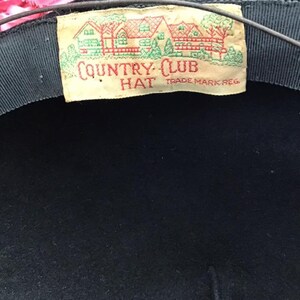 Puede incluir: Un sombrero de fieltro negro con una etiqueta blanca que dice "COUNTRY CLUB HAT TRADE MARK REG."