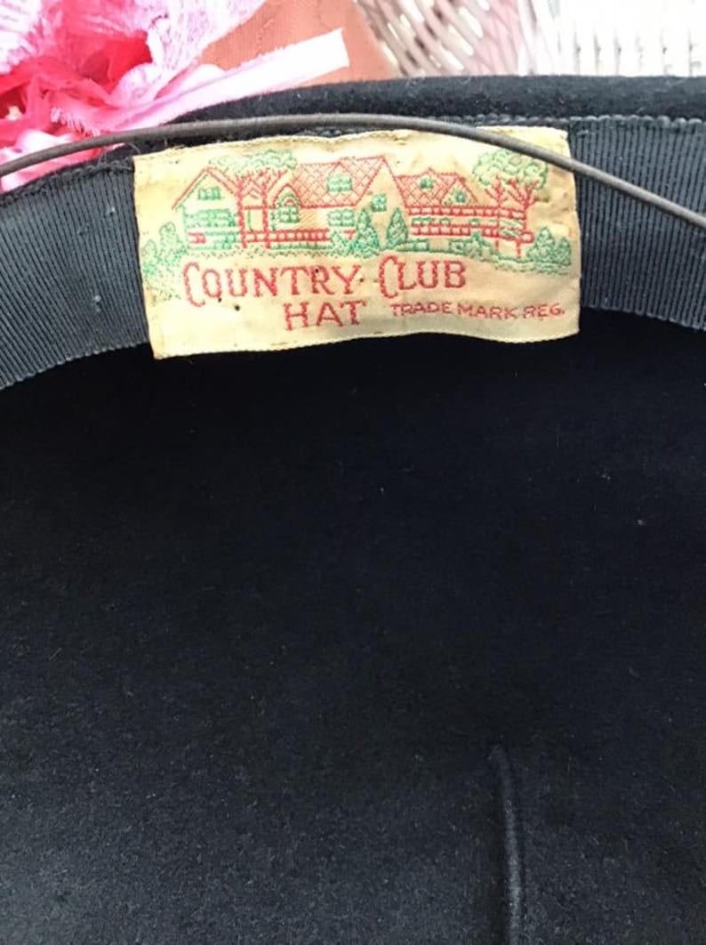 Puede incluir: Un sombrero de fieltro negro con una etiqueta blanca que dice "COUNTRY CLUB HAT TRADE MARK REG."