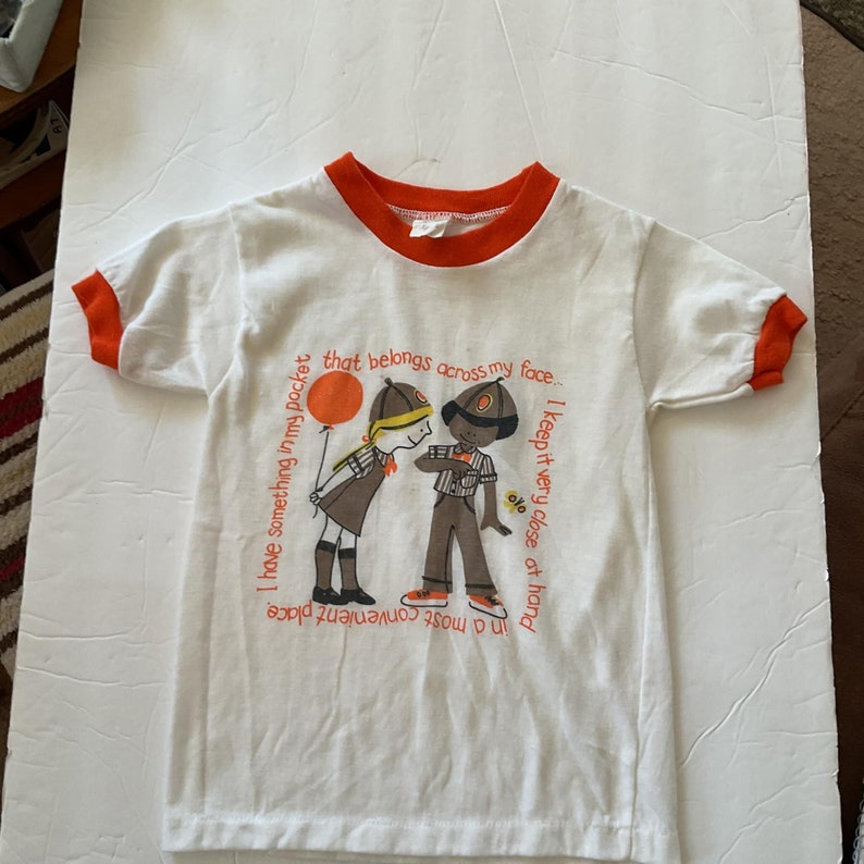 May include: White t-shirt with an orange ringer and a graphic of two children. The girl is holding a red balloon and the boy is holding a small yellow flower. The text on the shirt reads: "I have something in my pocket that belongs across my face... I keep it very close at hand in a most convenient place."