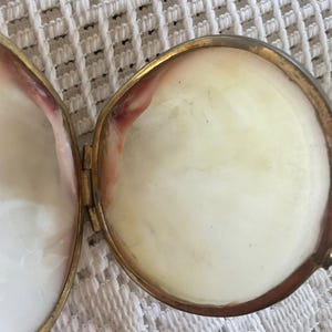 May include: Open, antique, gold-toned locket with a round shape. The locket is hinged and reveals a creamy white interior with a hint of pink and red. The locket is open, showing the inside of the piece.