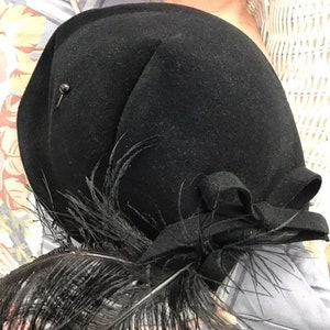 Puede incluir: Un sombrero de fieltro negro con una pluma negra y un lazo negro. El sombrero tiene un peque&ntilde;o alfiler negro en el lateral.