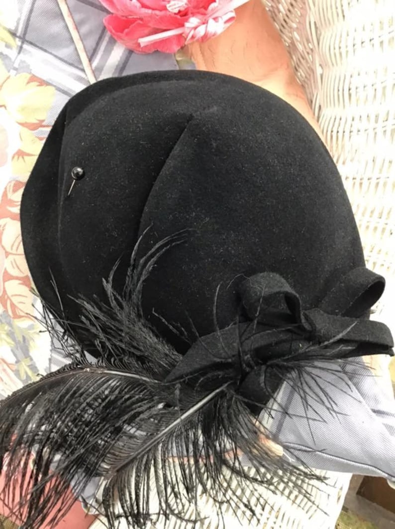 Puede incluir: Un sombrero de fieltro negro con una pluma negra y un lazo negro. El sombrero tiene un peque&ntilde;o alfiler negro en el lateral.