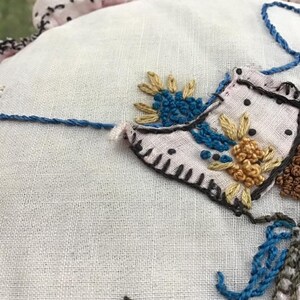 K&ouml;nnte beinhalten: Nahaufnahme eines wei&szlig;en Stoffes mit gestickten Blumen in Blaut&ouml;nen, Gelb und Braun. Der Stoff hat eine zarte Textur und ist von einem rosa und schwarz gepunkteten Besatz umgeben.