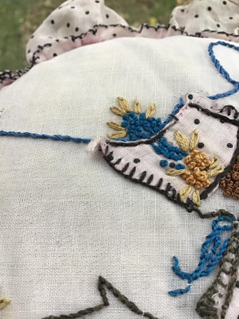 K&ouml;nnte beinhalten: Nahaufnahme eines wei&szlig;en Stoffes mit gestickten Blumen in Blaut&ouml;nen, Gelb und Braun. Der Stoff hat eine zarte Textur und ist von einem rosa und schwarz gepunkteten Besatz umgeben.