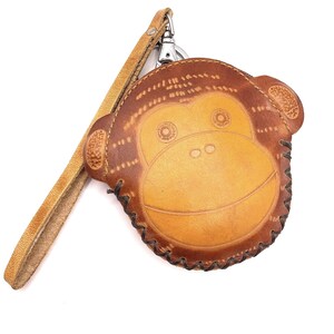 May include: A brown leather keychain shaped like a monkey's face. The keychain has a light brown face and darker brown details, including stitched features and a strap. The strap is a lighter shade of brown and is attached to a silver-colored clasp.