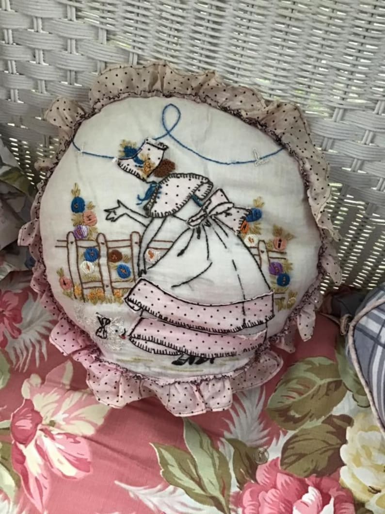 K&ouml;nnte beinhalten: Ein rundes, wei&szlig;es Kissen mit einem rosa-wei&szlig;en R&uuml;schenbesatz. Das Kissen ist mit einer Frau in einem rosa Kleid und einem blauen Hut bestickt, die in einem Garten mit einem Zaun und Blumen steht.
