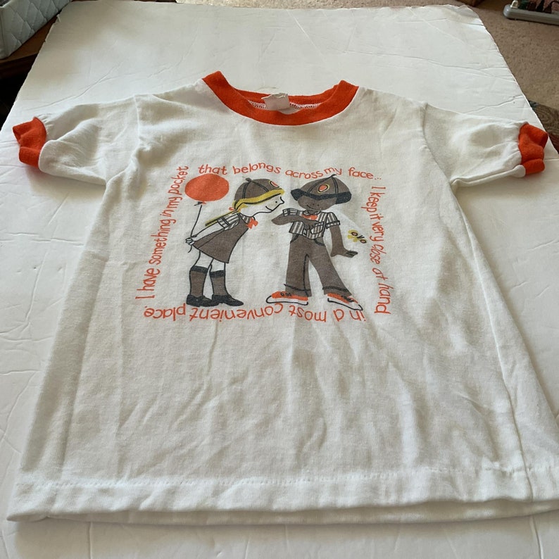 May include: White t-shirt with orange trim and a graphic of two children. The text on the shirt reads: "I have something in my pocket that belongs across my face. I keep it very close at hand in a most convenient place."