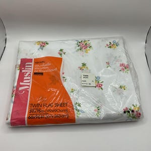 May include: A white twin flat sheet with a floral print in pink, yellow, and blue. The sheet is packaged in plastic and has a label that says "Muslin" and "no iron". The label also includes the thread count and fabric composition. The sheet is 39x75 inches (99x190 cm) and 66x96 inches (167x243 cm) after hemming.