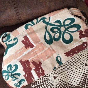 May include: A beige canvas tote bag with a green and brown abstract pattern. The bag has a single strap and is lying on a brown surface.