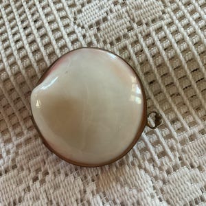 May include: A round, white and pink seashell-shaped trinket box with a brass-colored metal rim and a small handle. The box is sitting on a white, textured surface.