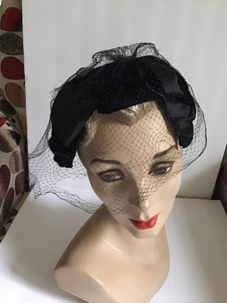Puede incluir: Un sombrero de terciopelo negro con velo de red negro. El sombrero tiene forma de diadema y tiene un lazo en un lado.