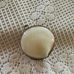 May include: A small, round, antique trinket box with a creamy white, slightly iridescent top. The box has a thin, bronze-colored metal frame and a small loop for hanging. The background is a white, open-weave fabric.