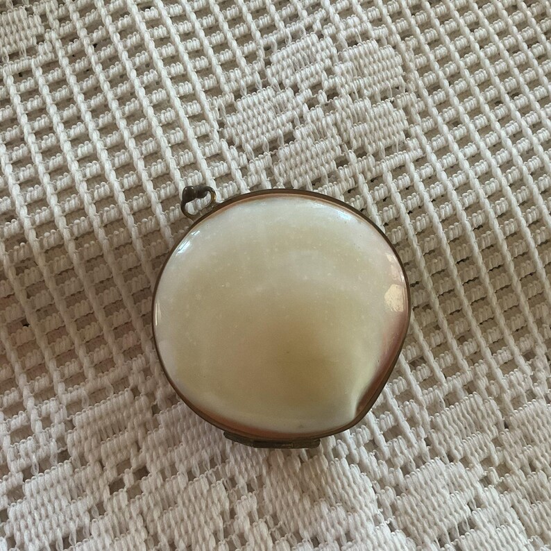 May include: A small, round, antique trinket box with a creamy white, slightly iridescent top. The box has a thin, bronze-colored metal frame and a small loop for hanging. The background is a white, open-weave fabric.