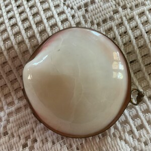 May include: A round, white and pink shell-shaped trinket box with a bronze-colored metal rim and clasp. The box has a glossy finish and is set against a white, textured background.