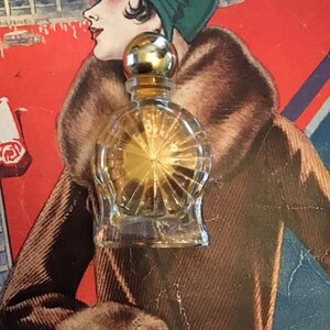 Puede incluir: Un peque&ntilde;o frasco de perfume de vidrio transparente con un cuerpo redondeado que presenta un patr&oacute;n de rayos de sol grabado. El frasco tiene un tap&oacute;n esf&eacute;rico dorado liso y contiene un l&iacute;quido dorado, sobre un fondo rojo.