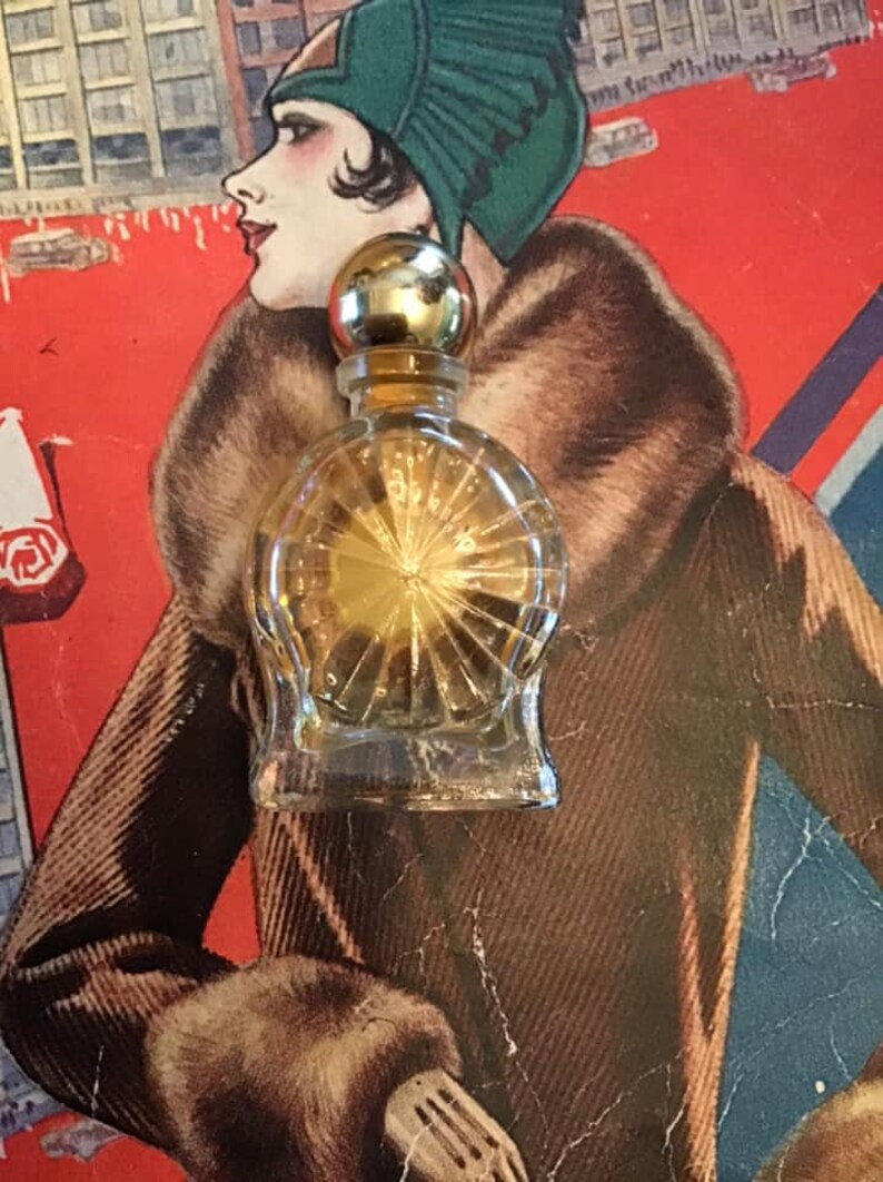 Puede incluir: Un peque&ntilde;o frasco de perfume de vidrio transparente con un cuerpo redondeado que presenta un patr&oacute;n de rayos de sol grabado. El frasco tiene un tap&oacute;n esf&eacute;rico dorado liso y contiene un l&iacute;quido dorado, sobre un fondo rojo.