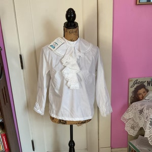 Könnte beinhalten: Weiße Bluse mit langen Ärmeln, Rüschenkragen und Spitzenbesatz. Die Bluse befindet sich auf einer Puppe und hat ein Etikett mit der Aufschrift "Princess Pearl".