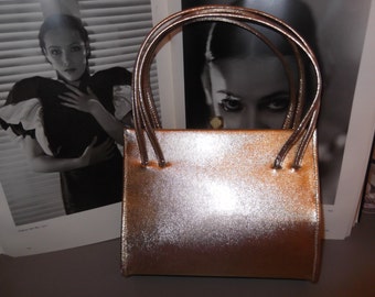 May include: A metallic gold clutch purse with a rectangular shape and two top handles. The purse has a smooth, shiny finish.