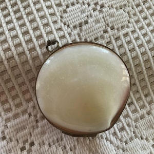 May include: A round, antique compact with a creamy white enamel top and a bronze-colored metal frame. The compact has a small loop for hanging and is set against a textured, off-white background.