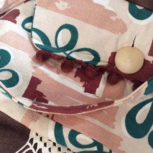 May include: A beige fabric bag with a flap closure and a brown pom-pom trim. The bag has a green and pink abstract pattern and a wooden button.