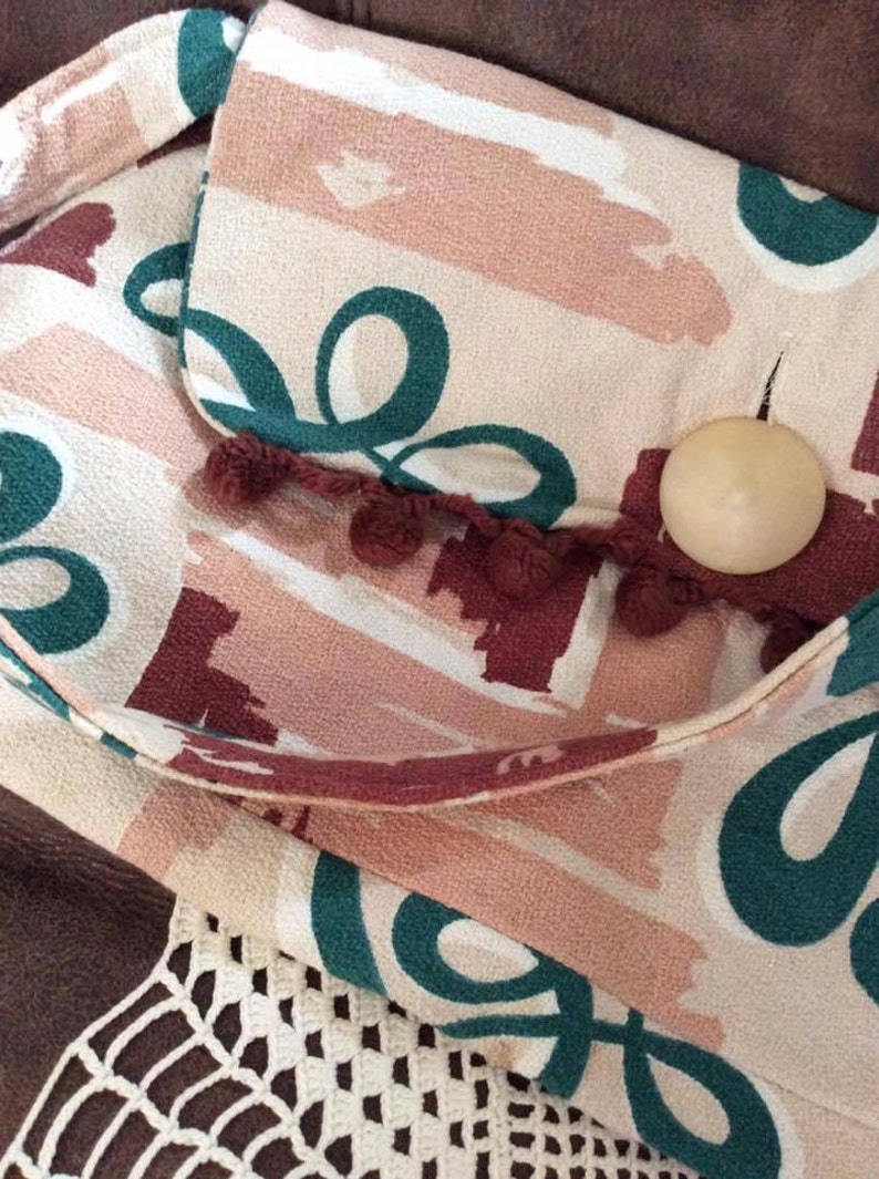 May include: A beige fabric bag with a flap closure and a brown pom-pom trim. The bag has a green and pink abstract pattern and a wooden button.