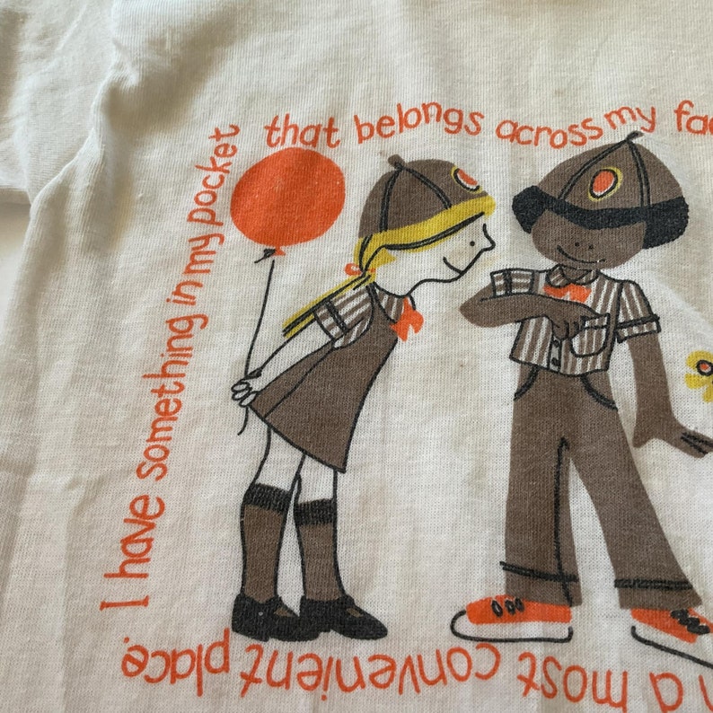 May include: A white t-shirt with a graphic of two children wearing brown uniforms. The girl is holding a red balloon and the boy is holding a flower. The text on the shirt reads "I have something in my pocket that belongs across my face."