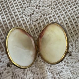 May include: An open, oval-shaped, hinged trinket box with a gold-colored metal frame. The interior is lined with a pearlescent material, with a hint of pinkish-red near the edges. The box is resting on a white, patterned surface.