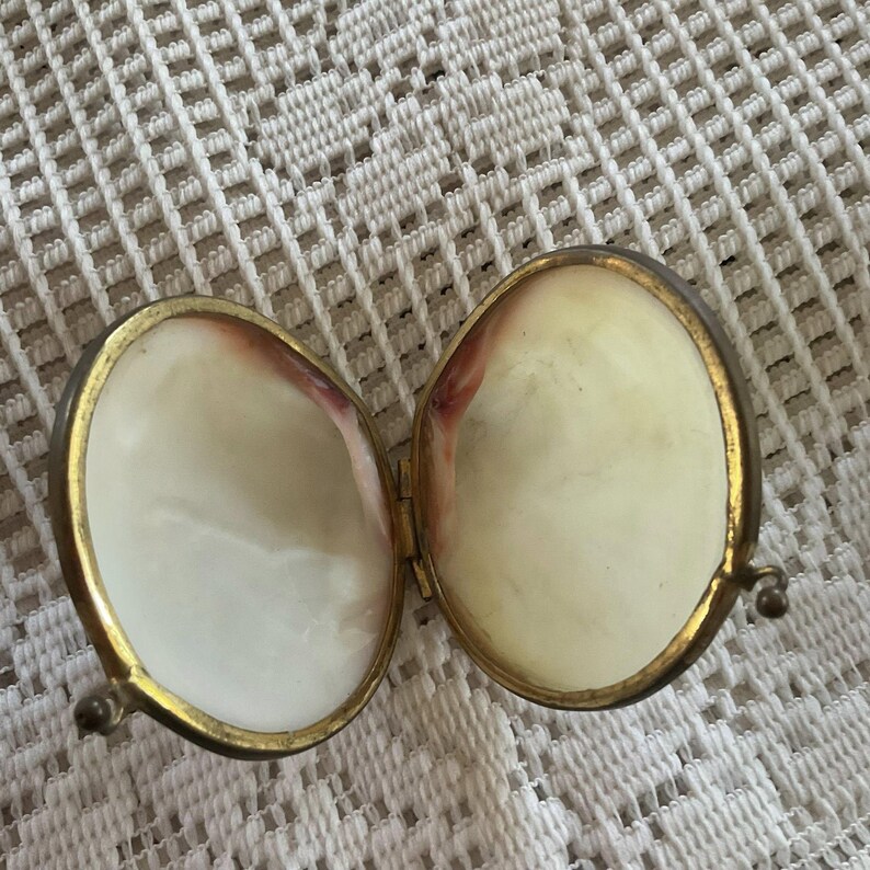 May include: An open, oval-shaped, hinged trinket box with a gold-colored metal frame. The interior is lined with a pearlescent material, with a hint of pinkish-red near the edges. The box is resting on a white, patterned surface.