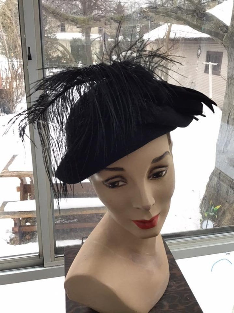 Puede incluir: Un sombrero de fieltro negro con una gran pluma negra. El sombrero est&aacute; colocado sobre una cabeza de maniqu&iacute;.