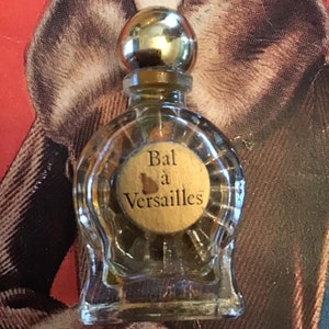 Puede incluir: Un frasco de perfume peque&ntilde;o de vidrio transparente con cuerpo redondo y tap&oacute;n esf&eacute;rico de color dorado. El frasco presenta una etiqueta circular beige en el frente con el texto Bal &agrave; Versailles impreso en negro. El vidrio tiene estr&iacute;as verticales.
