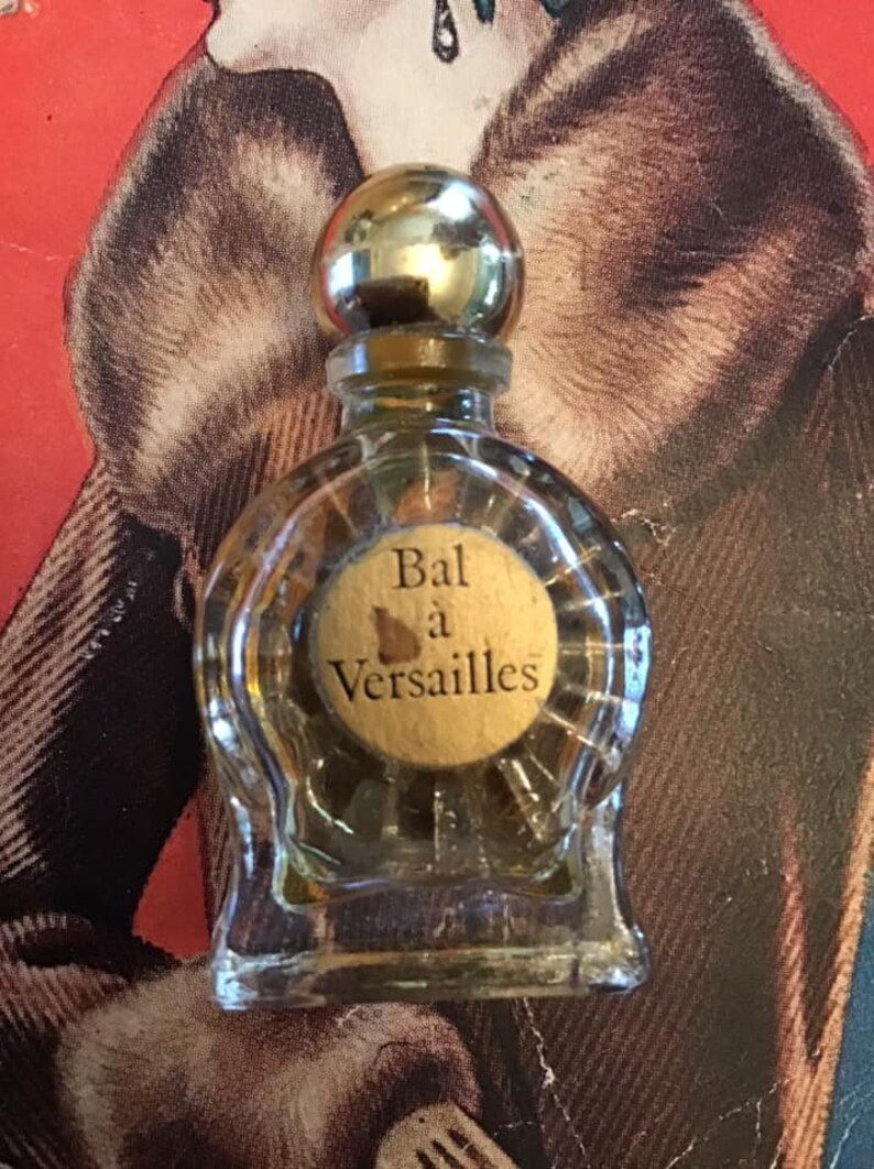 Puede incluir: Un frasco de perfume peque&ntilde;o de vidrio transparente con cuerpo redondo y tap&oacute;n esf&eacute;rico de color dorado. El frasco presenta una etiqueta circular beige en el frente con el texto Bal &agrave; Versailles impreso en negro. El vidrio tiene estr&iacute;as verticales.