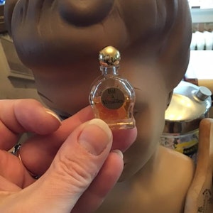 Puede incluir: Un frasco de perfume en miniatura de vidrio transparente con un tap&oacute;n esf&eacute;rico de tono dorado, que contiene un l&iacute;quido de color &aacute;mbar. La etiqueta frontal presenta el texto Bal a Versailles en letras negras sobre el cuerpo redondeado.